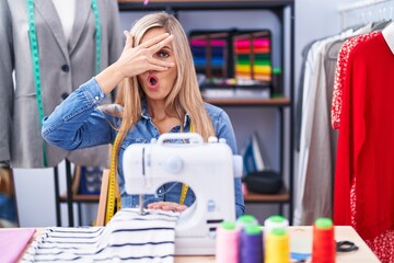 Blonde woman dressmaker designer using sew machine peeking in shock covering face and eyes with hand, looking through fingers with embarrassed expression.