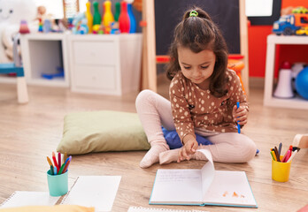 Adorable hispanic girl preschool student sitting on floor drawing on notebook at kindergarten