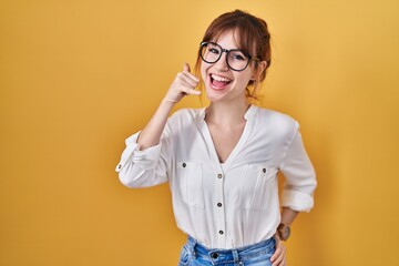 Young beautiful woman wearing casual shirt over yellow background smiling doing phone gesture with hand and fingers like talking on the telephone. communicating concepts.