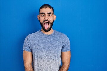 Middle east man with beard standing over blue background sticking tongue out happy with funny expression. emotion concept.