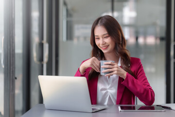 Pretty asian businesswoman sitting happily working on laptop and drinking coffee with bright smile in office.