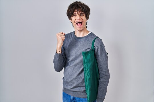 Young man wearing reusable bag screaming proud, celebrating victory and success very excited with raised arms