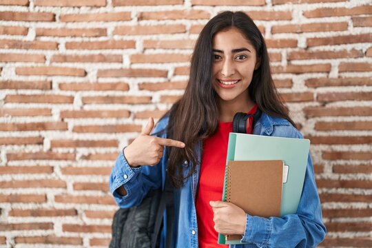 Young teenager girl wearing student backpack and holding books smiling happy pointing with hand and finger
