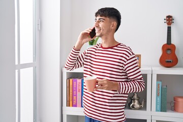 Young hispanic man talking on smartphone drinking coffee at bedroom