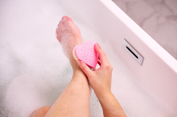 Woman scrubbing foot with sponge in bathtub - obrazy, fototapety, plakaty