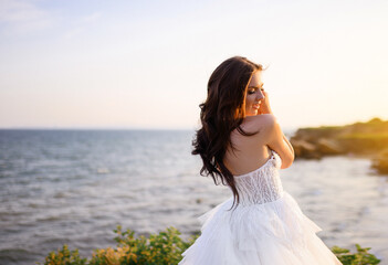 Fototapeta na wymiar Beautiful curly-haired bride model in wedding dress standing on top on sea background enjoying sun and nature. Charming girl in open sleeves dress.