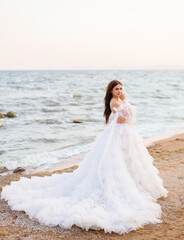 Charming bride woman model standing on sea beach in beautiful puffy wedding dress holding hand near attractive face, nature landscape on background.