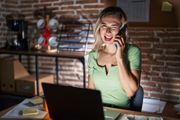 Young beautiful woman working at the office at night speaking on the phone winking looking at the...