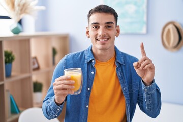 Handsome hispanic man drinking glass of orange juice surprised with an idea or question pointing finger with happy face, number one