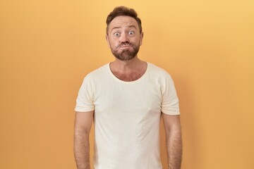 Middle age man with beard standing over yellow background puffing cheeks with funny face. mouth inflated with air, crazy expression. - obrazy, fototapety, plakaty