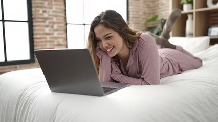 Naklejka premium Young beautiful hispanic woman using laptop lying on bed at bedroom