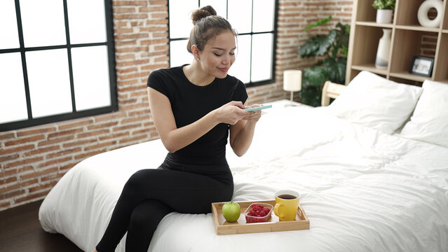 Young beautiful hispanic woman making photo to healthy breakfast by smartphone at bedroom