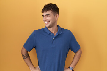 Young hispanic man standing over yellow background looking away to side with smile on face, natural expression. laughing confident.