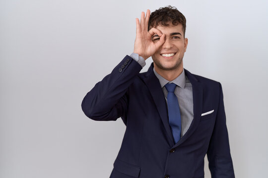 Young hispanic business man wearing suit and tie doing ok gesture with hand smiling, eye looking through fingers with happy face.