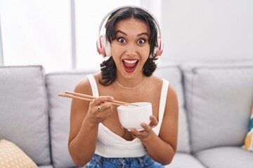 Young hispanic woman eating asian food using chopsticks celebrating crazy and amazed for success with open eyes screaming excited.