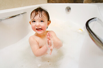 A happy child is playing with soap bubbles while sitting in a white home bathtub. Smiling toddler baby boy bathes in the bathroom. Kid aged one year six months