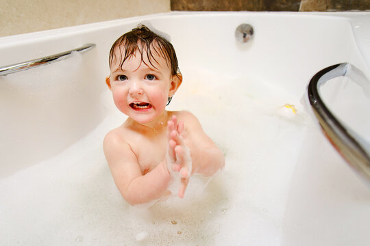 A happy child is playing with soap bubbles while sitting in a white home bathtub. Smiling toddler baby boy bathes in the bathroom. Kid aged one year six months