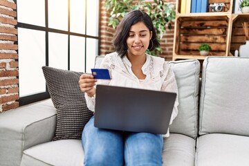 Fototapeta na wymiar Young latin woman using laptop and credit card sitting on sofa at home