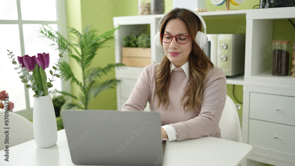 Sticker Young beautiful hispanic woman using laptop and headphones sitting on table at home