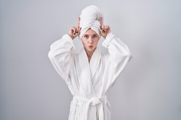 Blonde caucasian woman wearing bathrobe doing funny gesture with finger over head as bull horns