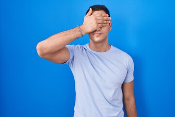 Young hispanic man standing over blue background covering eyes with hand, looking serious and sad....