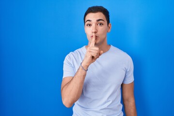Young hispanic man standing over blue background asking to be quiet with finger on lips. silence and secret concept.