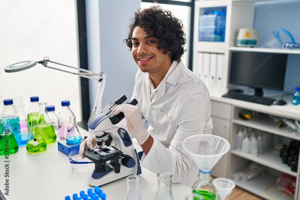 Sticker Young hispanic man wearing scientist uniform using microscope at laboratory