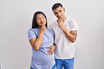 Young hispanic couple expecting a baby standing over background with hand on chin thinking about question, pensive expression. smiling with thoughtful face. doubt concept.