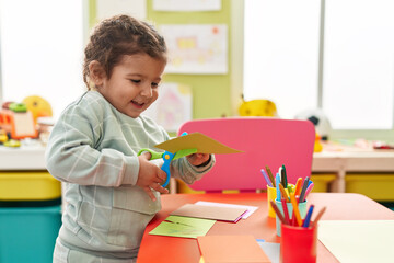 Adorable hispanic toddler student smiling confident cutting paper at kindergarten