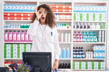 Hispanic woman with curly hair working at pharmacy drugstore doing ok gesture shocked with surprised face, eye looking through fingers. unbelieving expression.