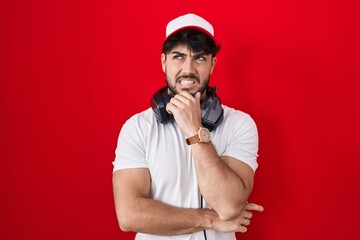 Hispanic man with beard wearing gamer hat and headphones thinking worried about a question, concerned and nervous with hand on chin