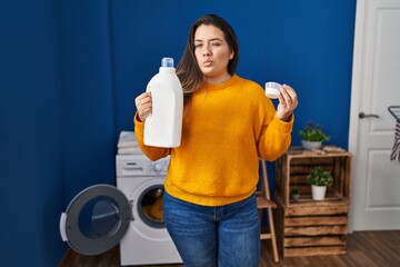 Young hispanic woman holding detergent bottle winking looking at the camera with sexy expression,...