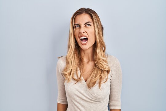 Young blonde woman standing over isolated background angry and mad screaming frustrated and furious, shouting with anger. rage and aggressive concept.