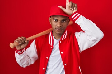Young hispanic man playing baseball holding bat making fun of people with fingers on forehead doing...