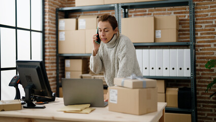 Middle age hispanic woman ecommerce business worker talking on the smartphone using laptop at storehouse office