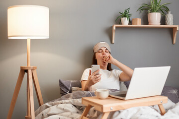 Portrait of sleepy beautiful woman wearing sleeping mask posing with on laptop while lying on bed, holding mobile phone, using smartphone for calling or for surfing internet. - obrazy, fototapety, plakaty