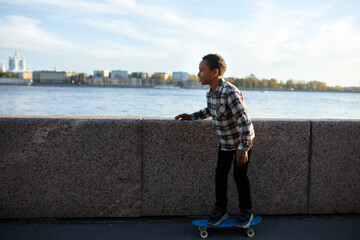 Side view of african american teen boy in plaid shirt riding skateboard along river embankment with picturesque cityscape on background, feeling insecure training on long board. Urban culture