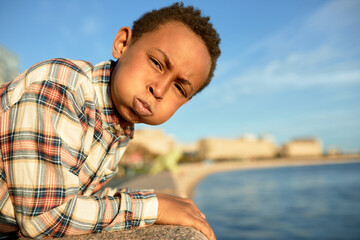 Displeased black boy of 13 blowing his cheeks looking at camera with angry facial expression, frowning, leaning on stoned fence of river embankment. Selective focus. Walking down city streets - obrazy, fototapety, plakaty