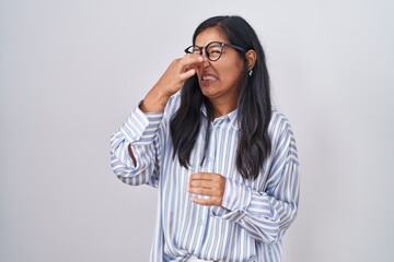 Young hispanic woman wearing glasses smelling something stinky and disgusting, intolerable smell,...