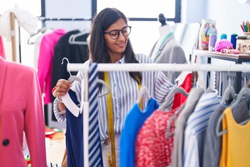 Young beautiful hispanic woman tailor smiling confident holding clothes on rack at clothing factory