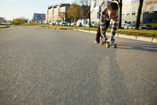 Low view image of cute active african american boy kid in plaid shirt accelerating on skateboard holding it with hands in his neighborhood, practicing and training outdoor. Urban culture