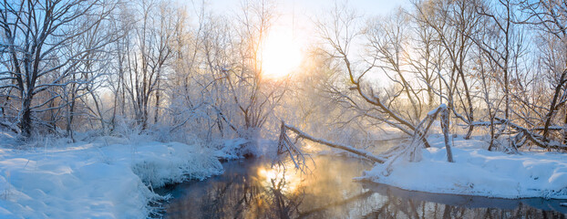 Winter sunny panoramic landscape with bare trees and forest river.Sun shining through the tree branches and fog rising from water.
