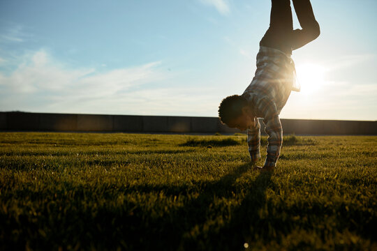 Outdoor image of active athletic sporty black boy of 13 standing on hands doing cartwheel in evening sun on green grass in public park. Urban culture, leisure activity. Copy space background