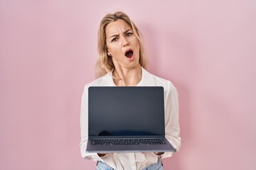 Young caucasian woman holding laptop showing screen in shock face, looking skeptical and sarcastic, surprised with open mouth