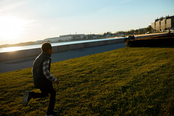 Fototapeta premium Side view of funny playful teenage boy running fast on green lawn on river embankment on warm spring evening. Urban leisure activity and healthy lifestyle. Happy carefree and active childhood