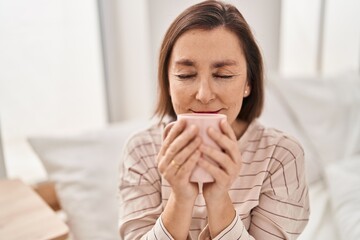 Middle age woman smelling coffee sitting on bed at bedroom