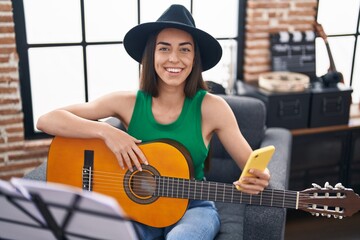 Young hispanic woman musician using smartphone playing classical guitar at music studio