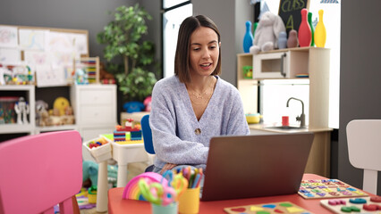 Fototapeta premium Young beautiful hispanic woman working as a teacher using laptop at kindergarten
