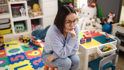 Young beautiful hispanic woman teacher sitting on table with stressed expression at kindergarten
