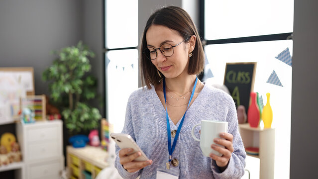 Young beautiful hispanic woman working as a teacher using smartphone drinking coffee at kindergarten - Powered by Adobe